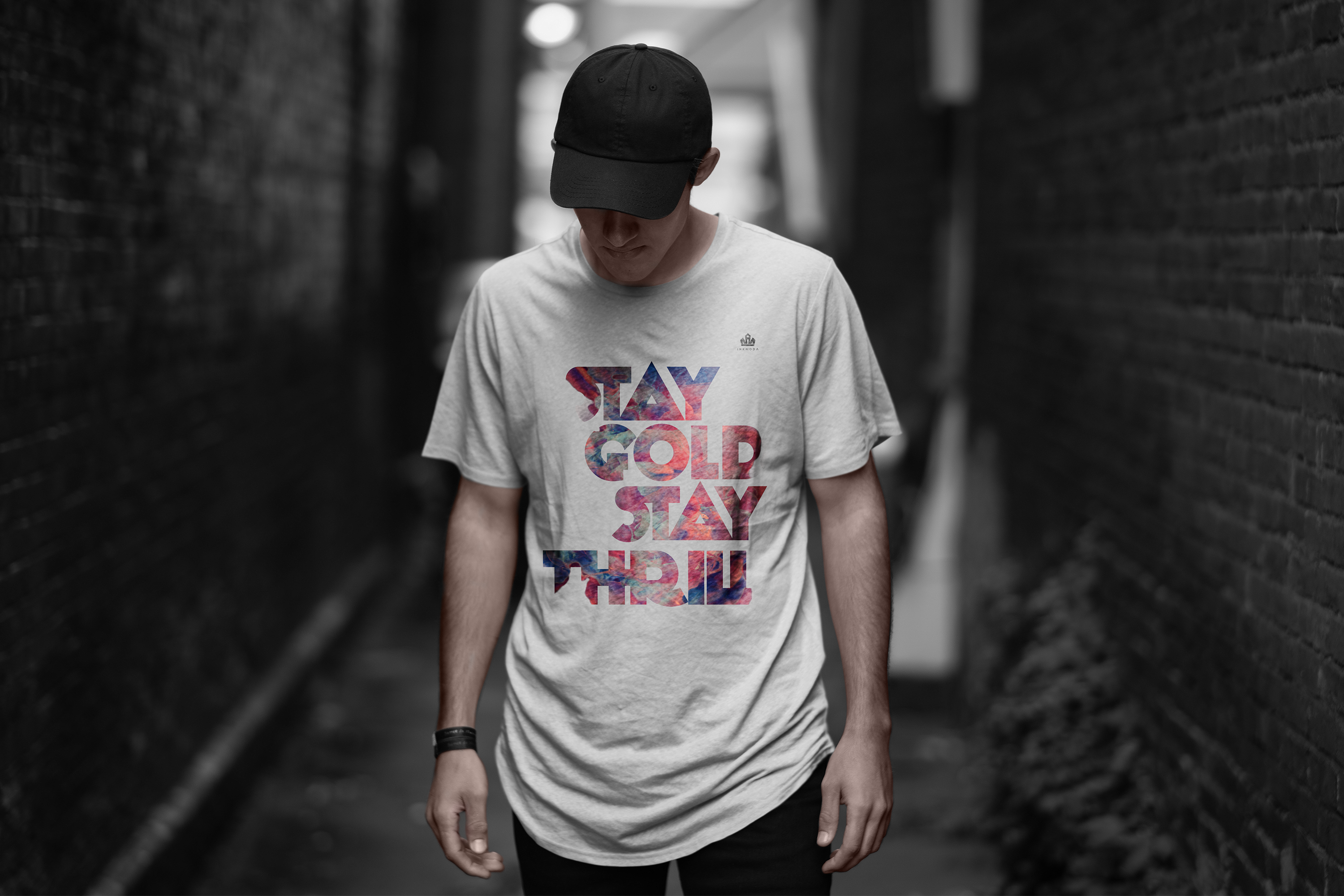 Male model in black cap and white long silhouette Inknoda tee in a dark alleyway wearing the STAY GOLD STAY THRILL typographic design with nebula watercolor gradient fill inside the letterforms